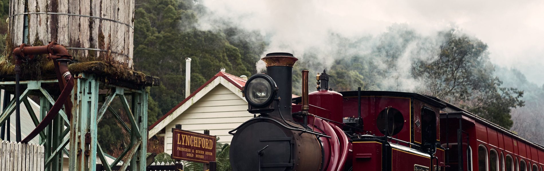 A steam locomotive waiting by the station's platform, a sign displaying that the train is stopped at Lynchford station. Nearby a water tank in the foreground, and steam bellowing from the loco's chimney.
