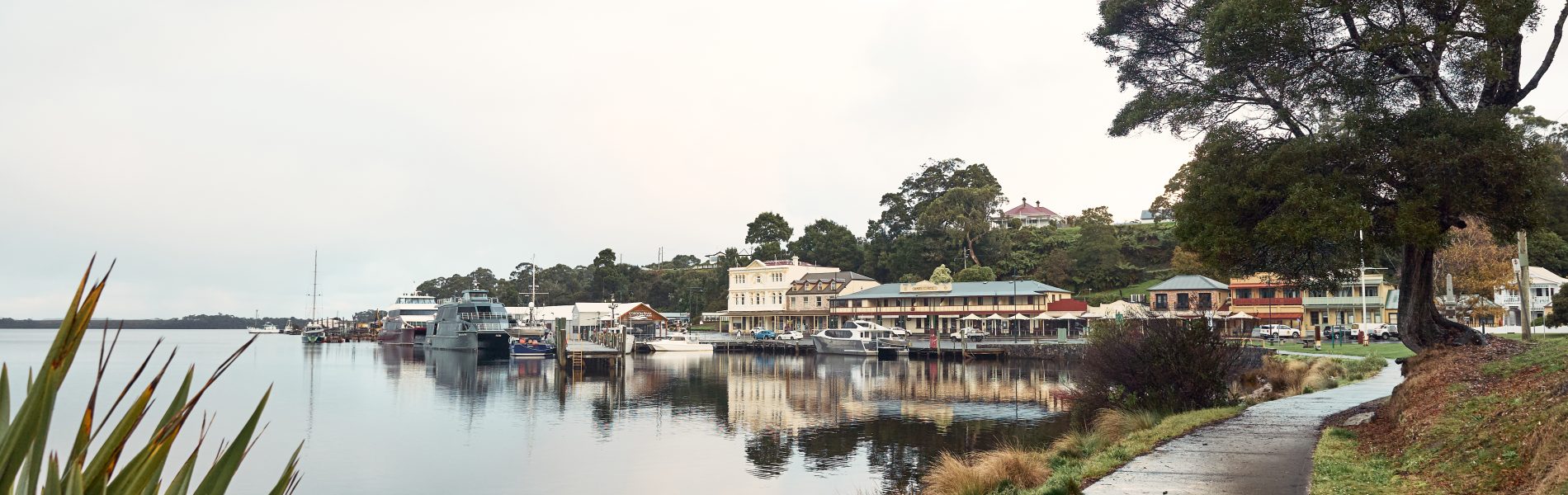 An image of the Strahan waterfront with a walking track to the right and the Macquarie Harbour to the left. Image also shows the main township of Strahan in the background along with boats that are docked.